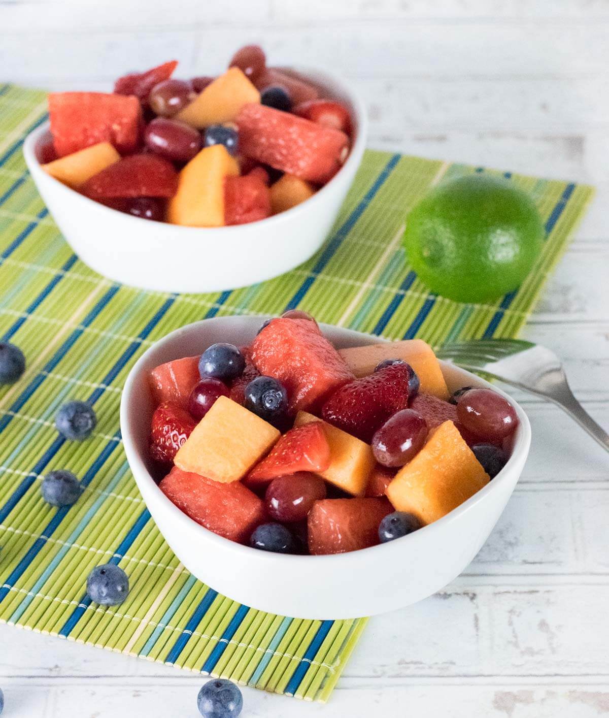 Two bowls of fruit salad with fresh lime and blueberries alongside.