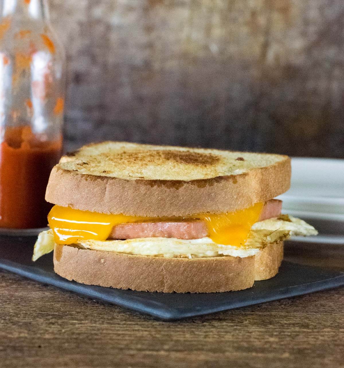 Spam Sandwich on black tray with hot sauce bottle behind.