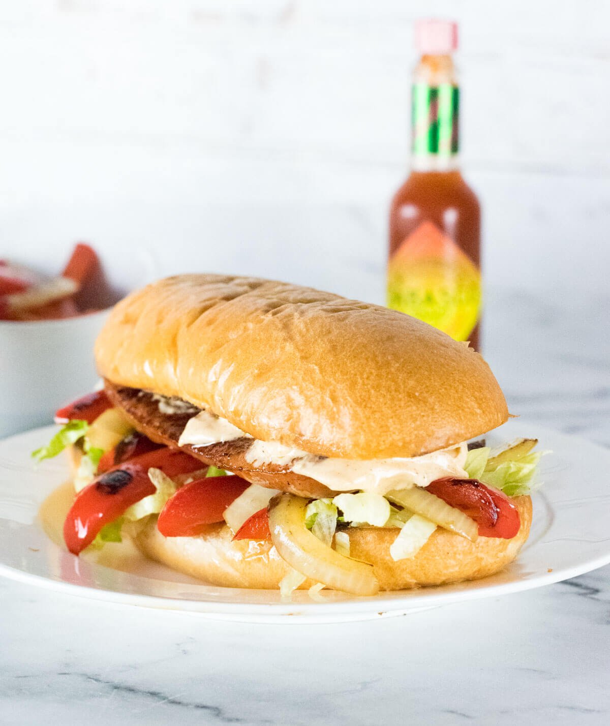 Andouille Sausage Sandwich sitting on a white plate with Tabasco sauce behind it.