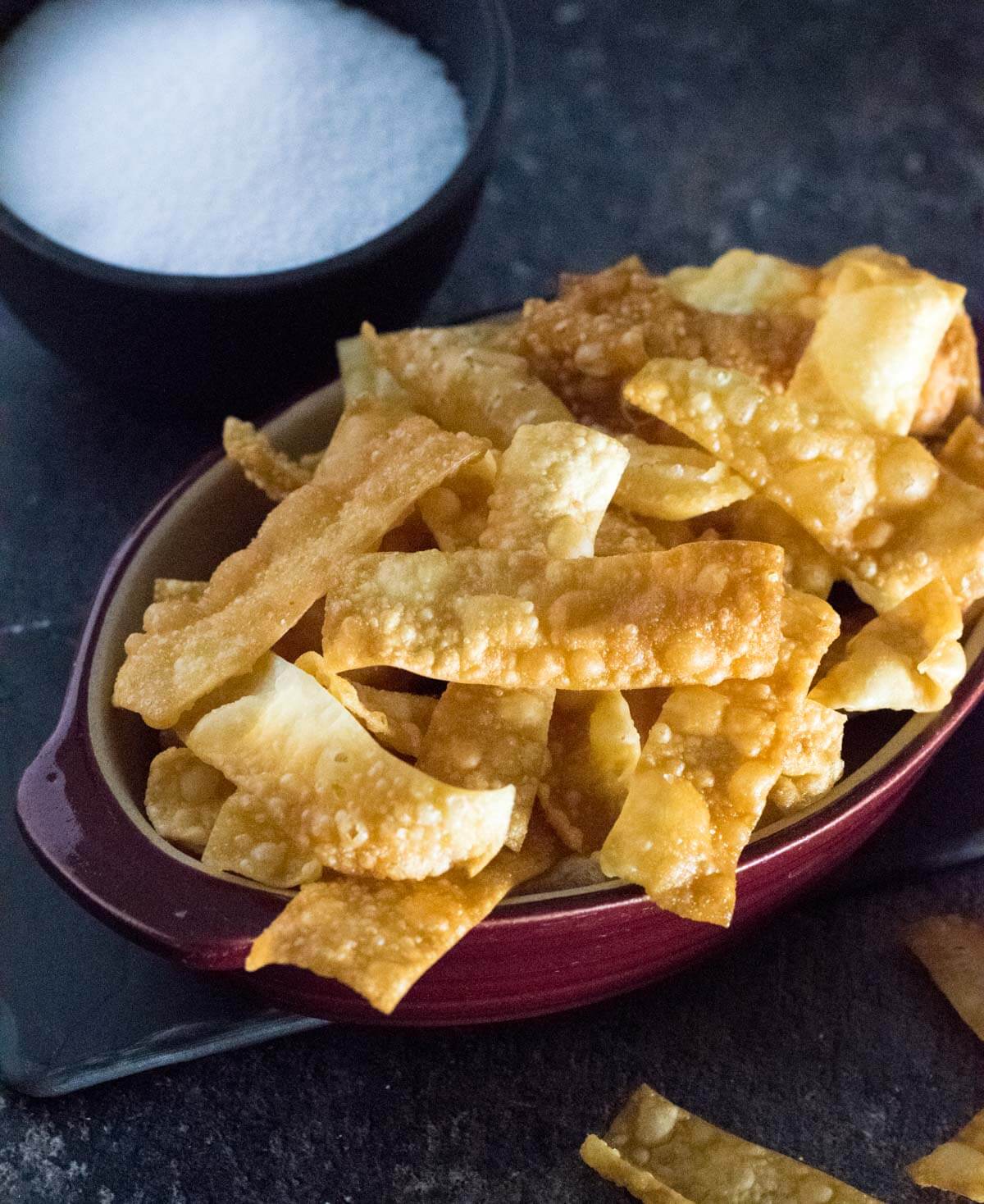 Fried wonton chips shown close up in red dish with black background.