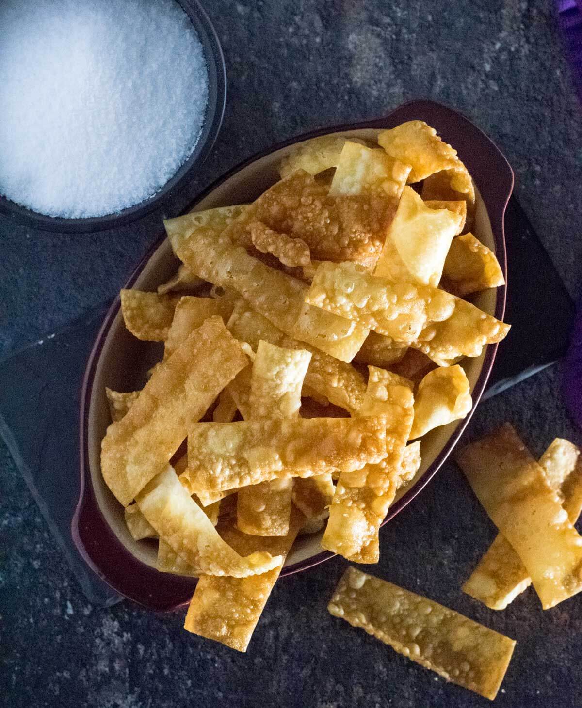 Red bowl of fried wonton strips with salt dish alongside.