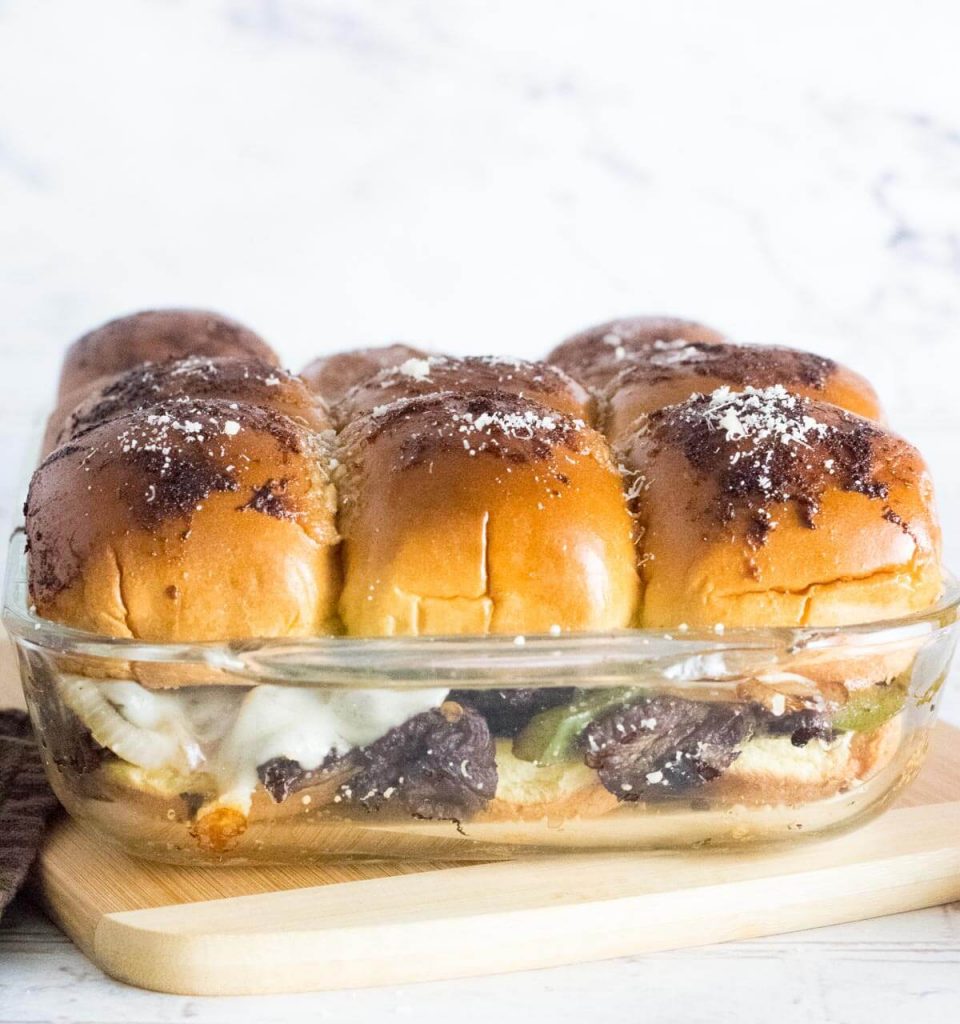 Philly cheesesteak sliders in a baking dish sitting on cutting board.