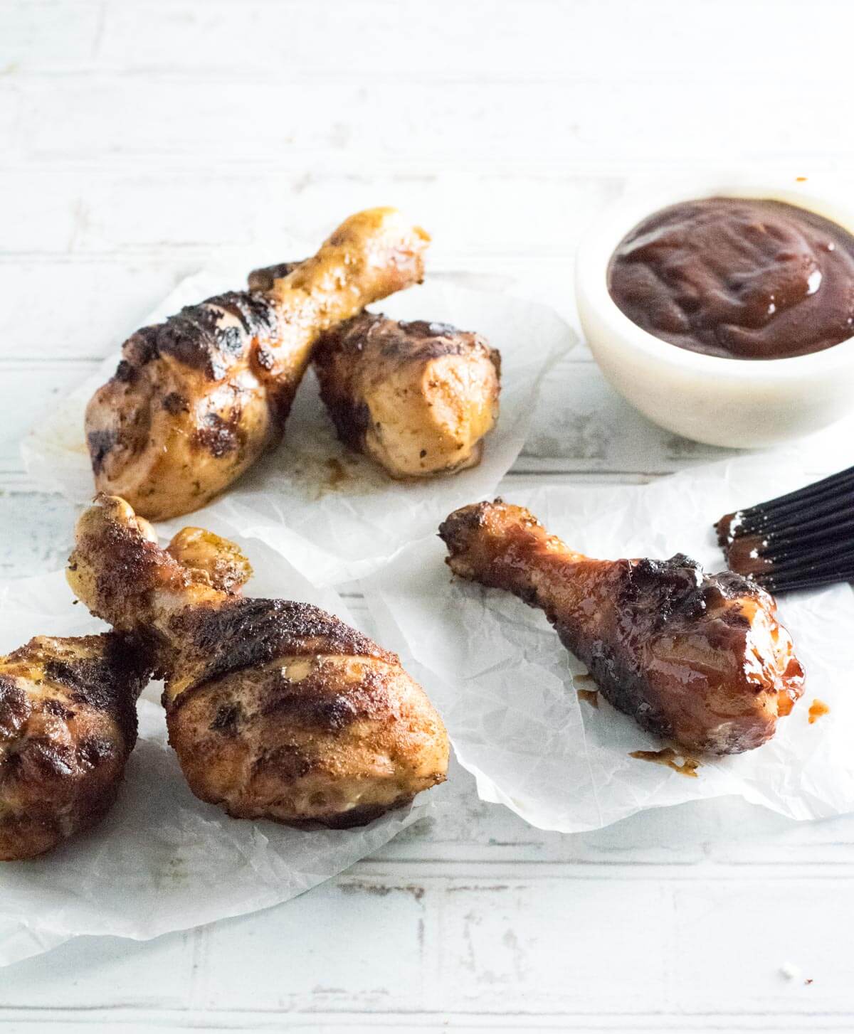 Grilled chicken drumsticks on wax paper with BBQ sauce in bowl.